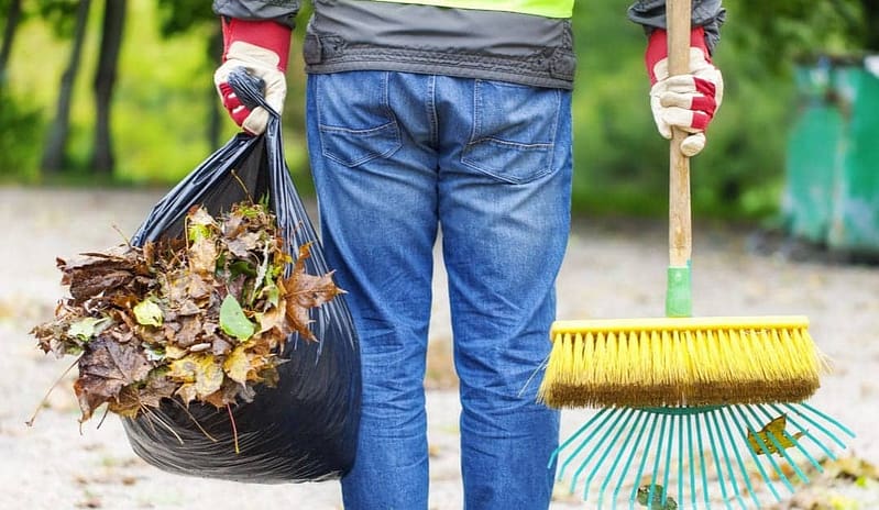 A man holding a bag of raked leaves, a rake, and a broom after cleaning up the yard.